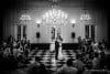 A bride and groom share their first dance in an elegant ballroom with chandeliers. Guests are seated around the checkered floor, watching the couple. The room has tall windows and ambient lighting, creating a romantic atmosphere.