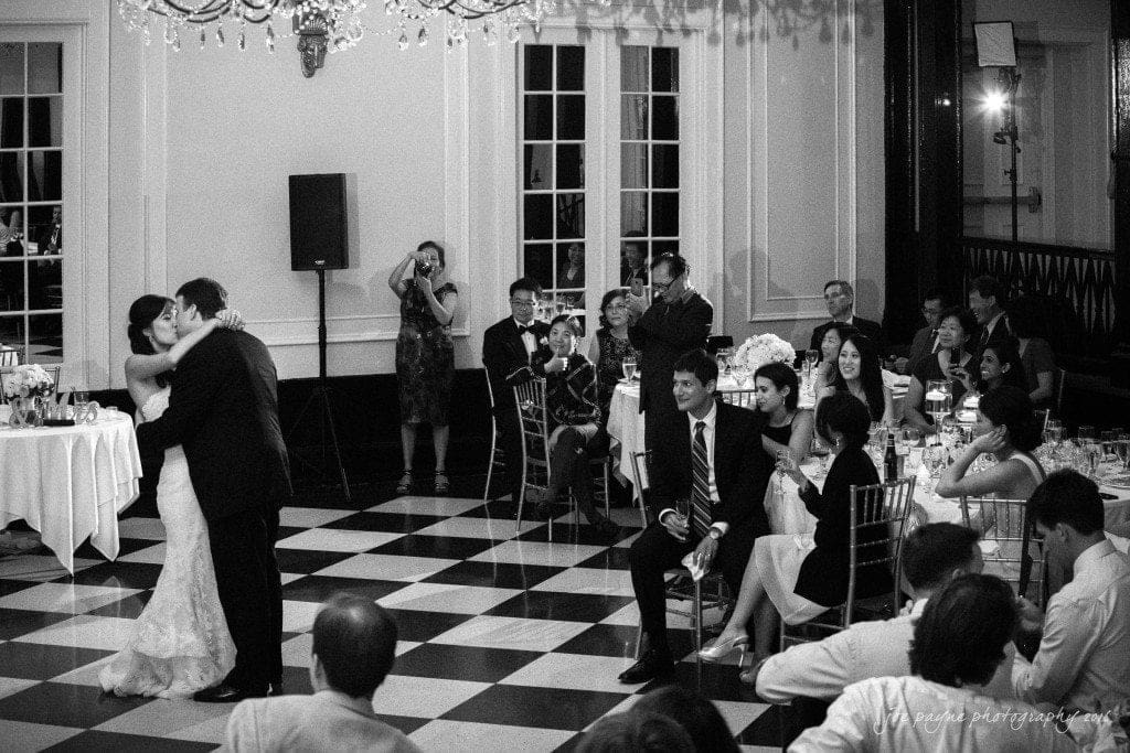 A bride and groom share their first dance in an elegant ballroom with black and white checkered floors. Guests watch and smile at the couple from their seats under a sparkling chandelier.