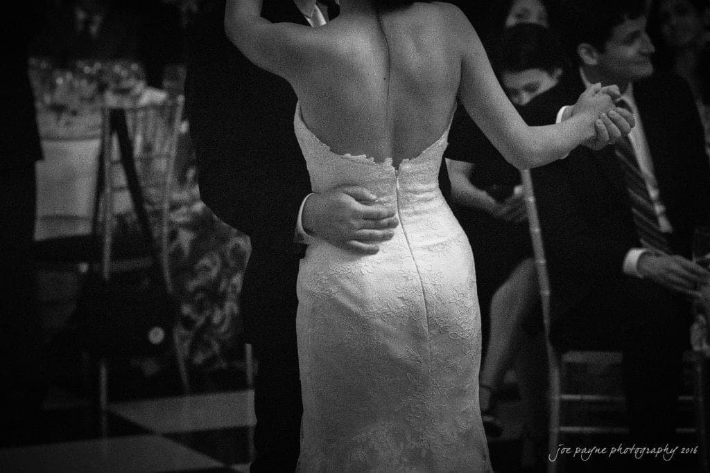 A groom and bride dance closely at their wedding reception, surrounded by guests seated at tables. The brides lace gown is visible as they share an intimate moment on the dance floor. The atmosphere is elegant and celebratory.