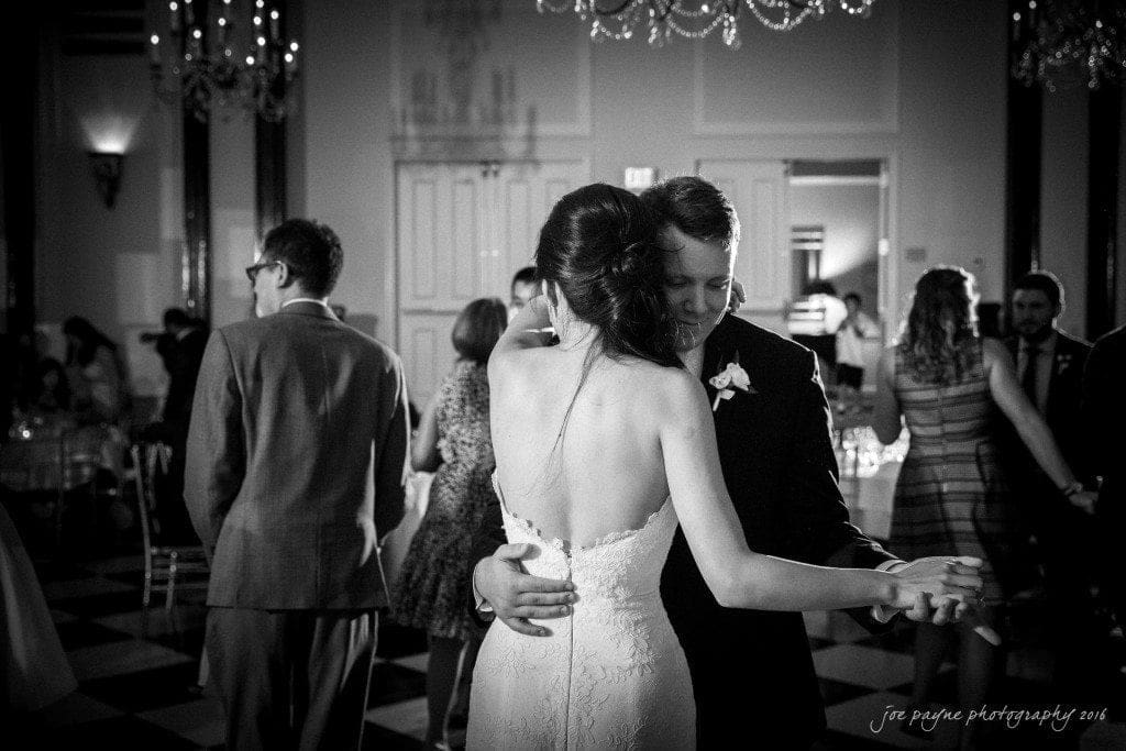 A bride and groom share a slow dance at their wedding reception. The bride wears a strapless gown, and the groom is in a suit. They are surrounded by guests in a dimly lit, elegant venue with chandeliers and a checkered floor.