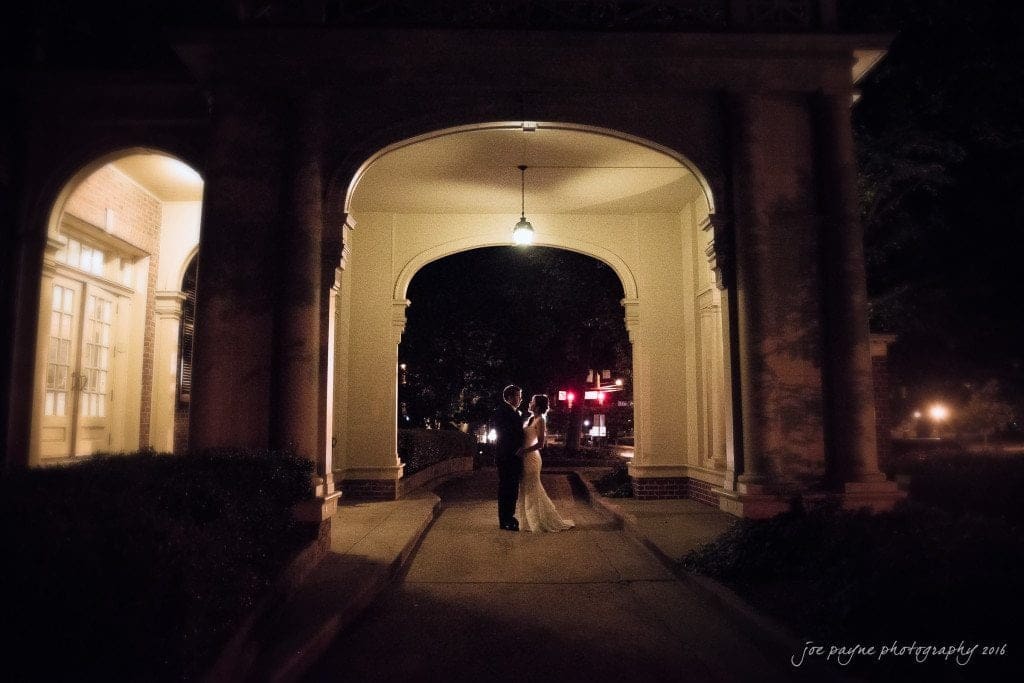 A couple stands embraced under a warmly lit archway at night. The soft, ambient light creates a romantic atmosphere as they share an intimate moment in a serene setting. The background is softly blurred, emphasizing their connection.