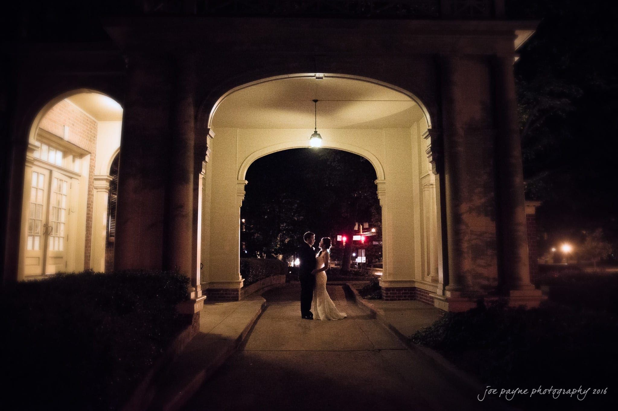 A couple stands embraced under a warmly lit archway at night. The soft, ambient light creates a romantic atmosphere as they share an intimate moment in a serene setting. The background is softly blurred, emphasizing their connection.