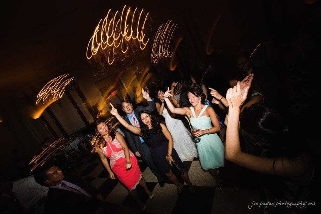 A group of people dance energetically on a checkered floor under swirling lights at a party. They are dressed in formal attire and appear to be enjoying themselves.