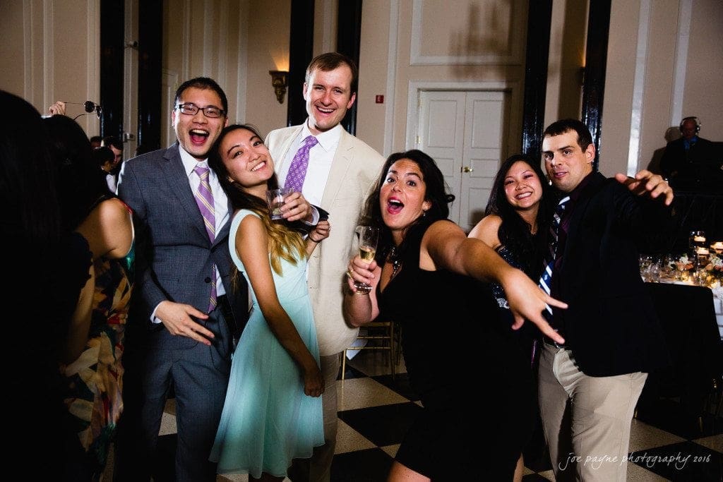 A group of six people in formal attire are joyfully posing and smiling for the camera at an indoor event with black and white checkered flooring. Some are holding drinks, and the lighting is warm and festive.