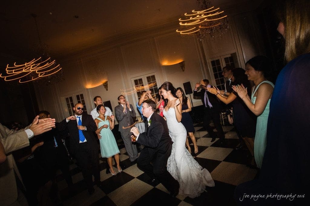 A joyful wedding reception with a bride and groom dancing at the center. The groom is kneeling playfully, surrounded by cheering guests on a checkered floor. Elegant chandeliers hang overhead, casting warm light.