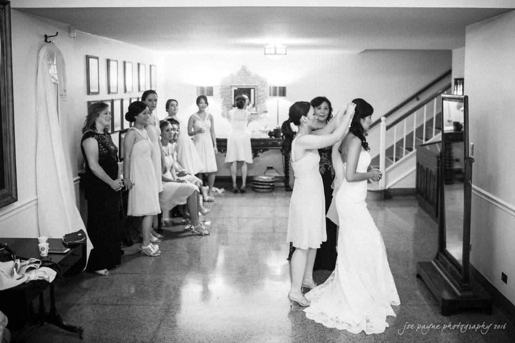 A bride stands in a room as a woman adjusts her veil. Several bridesmaids in dresses watch and smile. The setting is spacious with a staircase and framed pictures on the walls. The photo is in black and white.