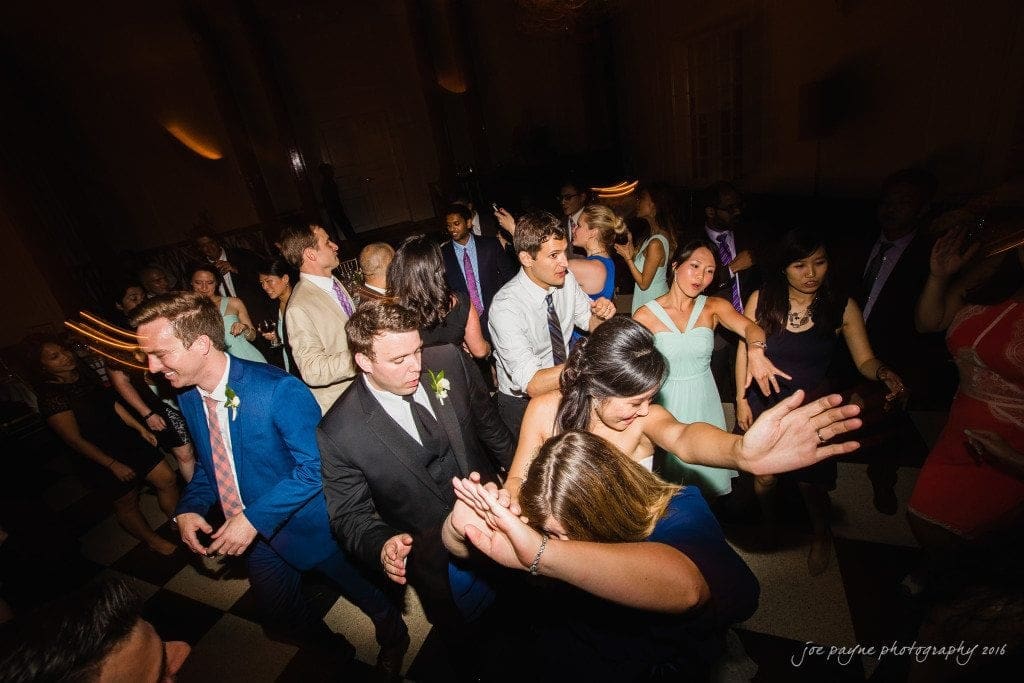 A lively group of people dancing at an indoor event. They are dressed in formal attire and appear to be enjoying themselves on a dimly lit dance floor.