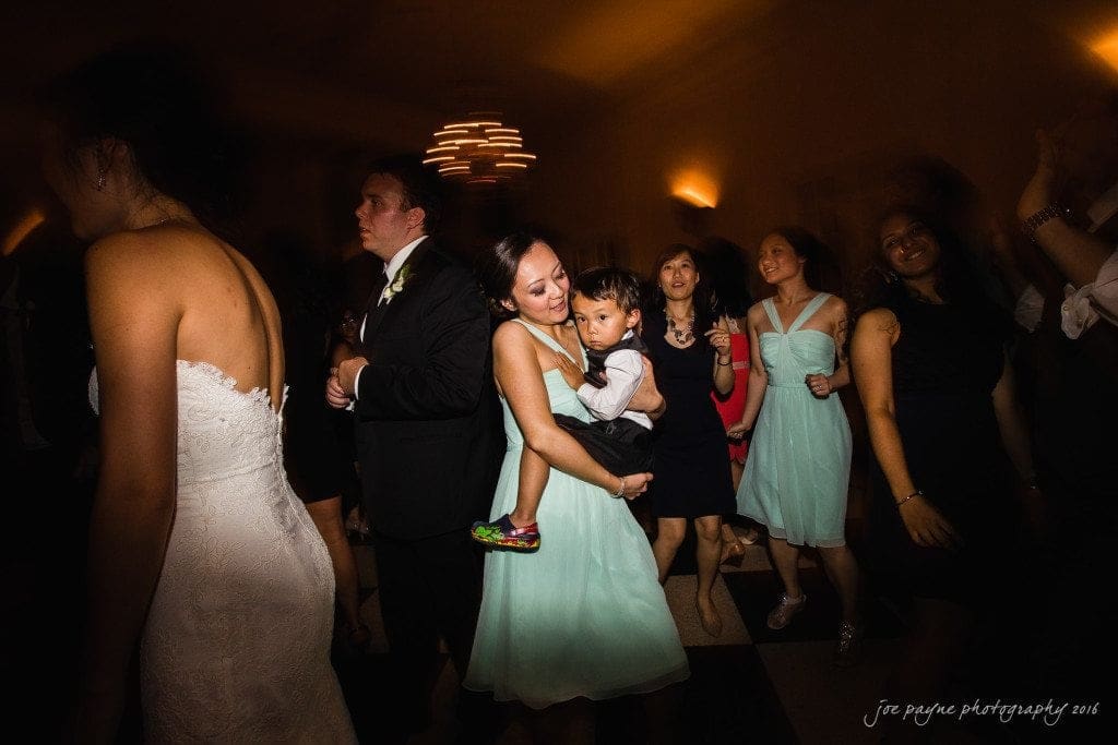 A woman in a light blue dress holds a child on a lively dance floor. Surrounding them are people in formal attire, dancing and enjoying the event. A chandelier softly illuminates the scene.