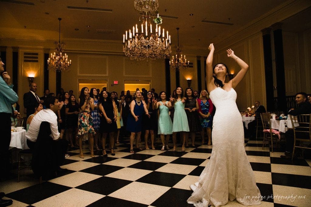 A bride in a white gown stands in a ballroom with checkered floors, preparing to throw her bouquet. Guests, dressed in various colors, eagerly watch in the background. Chandeliers hang from the ceiling, illuminating the elegant setting.