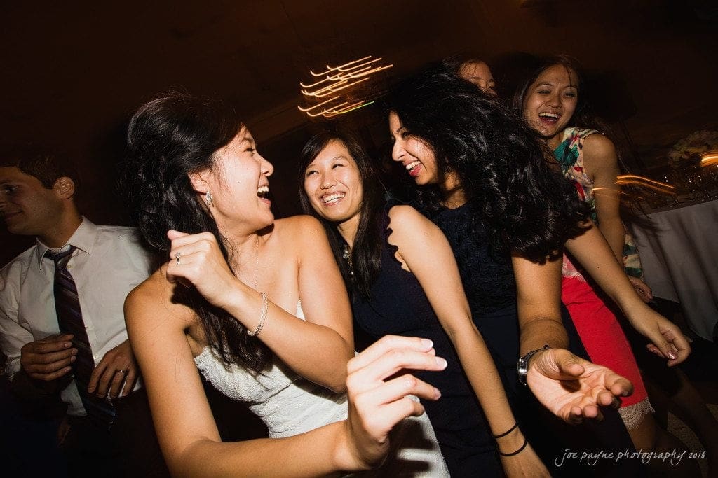 A joyous group of people dance closely together at a celebration. They are smiling and laughing, capturing a moment of fun and togetherness. The festive atmosphere is enhanced by dim lighting and blurred motion of festive lights in the background.