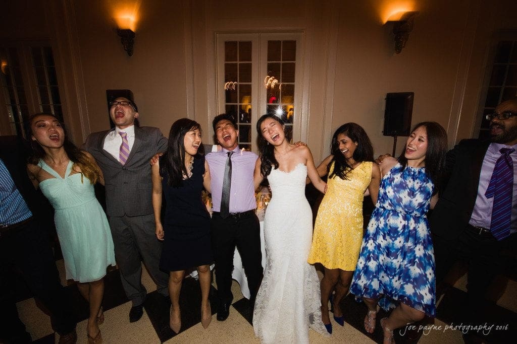 A group of people in formal attire stand closely together, laughing and having fun. They appear to be at a celebratory event, possibly a wedding, with the bride in a white dress at the center. The atmosphere is lively and joyful.