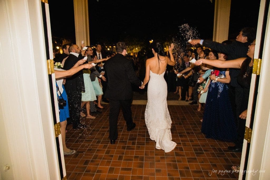 A newlywed couple, with the bride in a white dress and the groom in a black suit, walks down a hall as guests on either side throw rice and celebrate. The atmosphere is festive and joyful, highlighted by warm lighting.