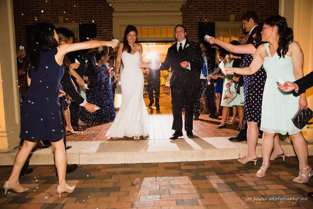 A bride and groom exit a venue surrounded by guests celebrating and tossing confetti. The bride is wearing a white wedding dress, and the groom is in a black suit. The scene is festive and joyful.