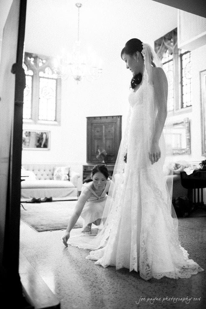 Bride in a long, elegant lace gown stands in a bright room with large windows and a chandelier. A bridesmaid adjusts the train of the dress. The room is decorated with plush furniture and framed pictures.