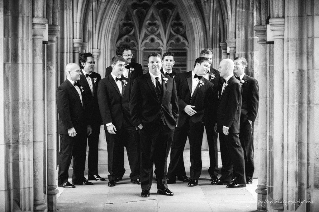 A group of groomsmen in black tuxedos stand in an ornate, arched hallway. They are smiling and conversing, with the groom at the center. The setting is elegant, featuring intricate stonework.