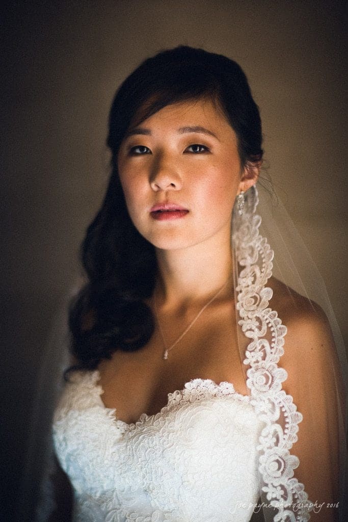 A bride wearing a lace-trimmed veil and elegant white wedding dress, with subtle makeup and pearl jewelry, stands against a dark background, gazing softly at the camera.