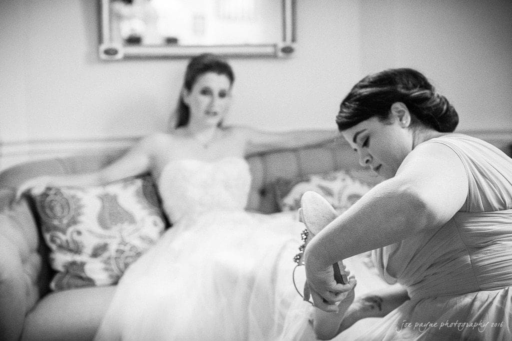duke chapel & carolina inn wedding - sidney & andrew A bride in a white dress sits on a couch looking on as a woman kneels in front of her, helping her put on a shoe. The room is softly lit, creating a calm atmosphere.