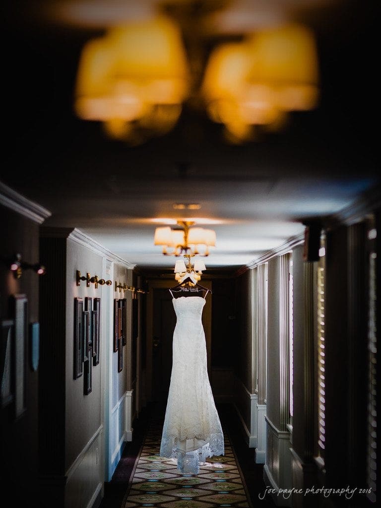 A white wedding dress hangs from a chandelier in a dimly lit hallway. Elegant wall sconces line the walls, and framed pictures are visible. Soft, natural light filters through the window blinds, highlighting the intricate details of the gown.