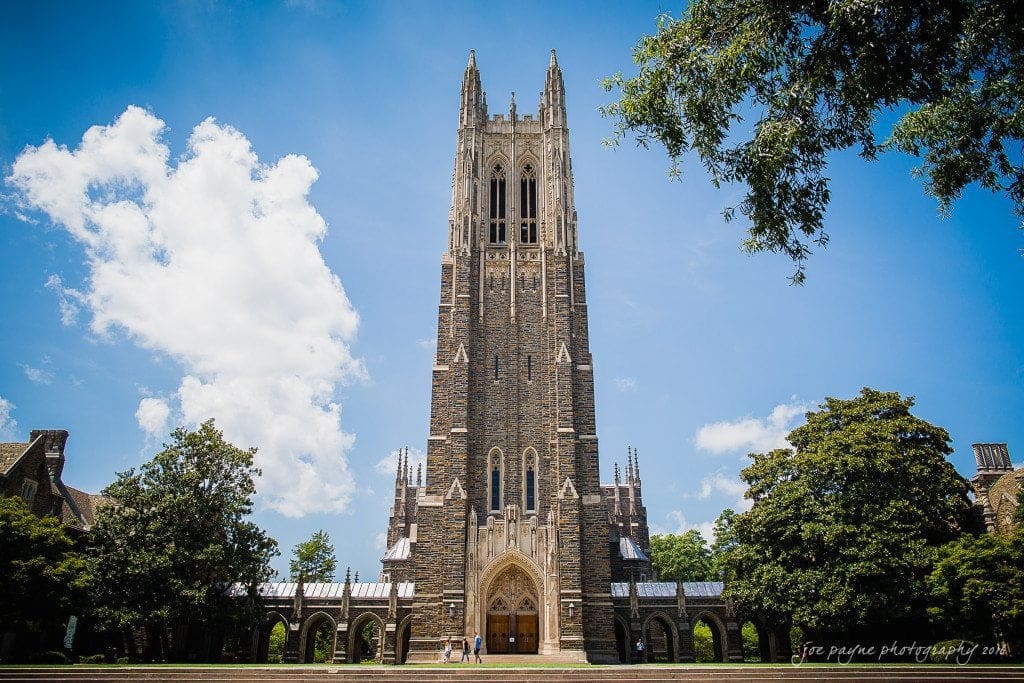 duke chapel & carolina inn wedding - sidney & andrew A grand stone chapel with a tall tower under a blue sky, surrounded by trees. People stand near the entrance, emphasizing the structures height and majesty.