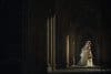 A bride in a long, flowing veil and gown stands in a sunlit archway, holding a bouquet. The architectural details and shadows create a dramatic atmosphere.