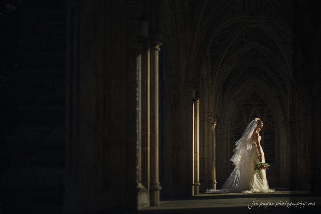 duke chapel & carolina inn wedding - sidney & andrew A bride in a long, flowing veil and gown stands in a sunlit archway, holding a bouquet. The architectural details and shadows create a dramatic atmosphere.