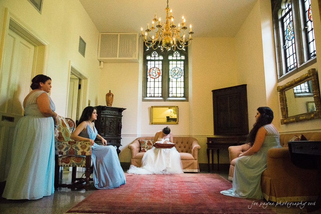 duke chapel & carolina inn wedding - sidney & andrew A bride in a white dress sits reading on a couch in an elegantly decorated room with stained glass windows. Three bridesmaids in light blue dresses sit and stand around her, all engaged in quiet conversation. A chandelier hangs above them.