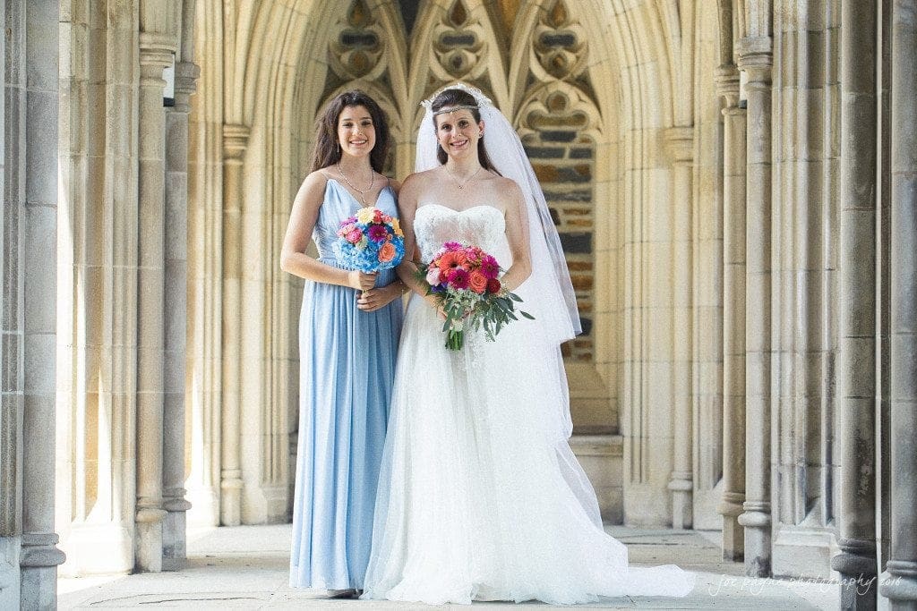 duke chapel & carolina inn wedding - sidney & andrew Two women stand in a stone archway. One wears a white wedding gown and veil, holding a bouquet of red and pink flowers. The other in a light blue dress holds a bouquet of bright pink flowers. The setting is elegant and sunlit.