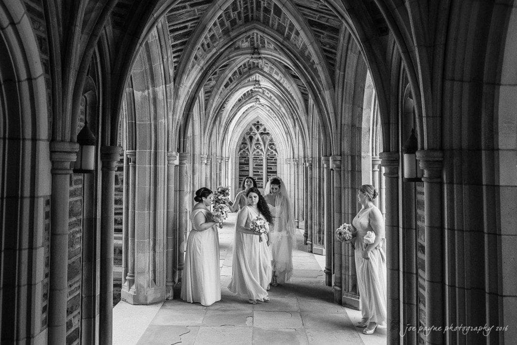 duke chapel & carolina inn wedding - sidney & andrew A black and white photo of five bridesmaids in long dresses standing in a stone archway. They appear to be talking and holding bouquets, surrounded by gothic-style architecture.