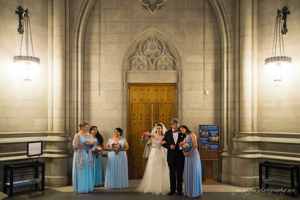 duke chapel & carolina inn wedding - sidney & andrew A bride and groom stand in the center of a stone hall with wooden doors, surrounded by six bridesmaids in blue dresses holding bouquets. The setting features ornate architecture and hanging lamps.