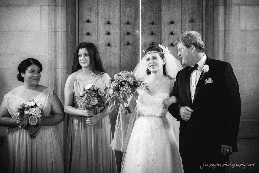 duke chapel & carolina inn wedding - sidney & andrew A bride in a wedding gown, holding a bouquet and standing arm-in-arm with a man in a tuxedo. Two bridesmaids in elegant dresses stand beside them, each holding a bouquet. They are all smiling. The setting appears to be an ornate indoor venue.