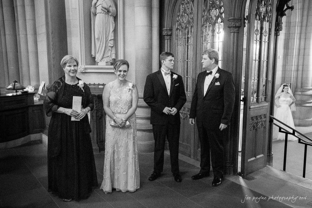 duke chapel & carolina inn wedding - sidney & andrew Four formally dressed adults stand inside a church entrance, chatting. Two women wear long dresses, and two men are in tuxedos. A bride in the background, partially visible, is walking up steps. The setting is elegant and decorated.