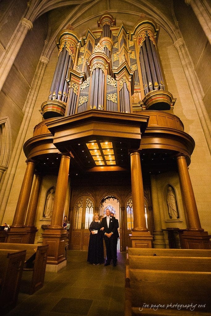duke chapel & carolina inn wedding - sidney & andrew A couple stands in the aisle of a grand cathedral, beneath a large, ornate pipe organ. The cathedral features tall columns and intricate architectural details. The atmosphere is warm and reverent.