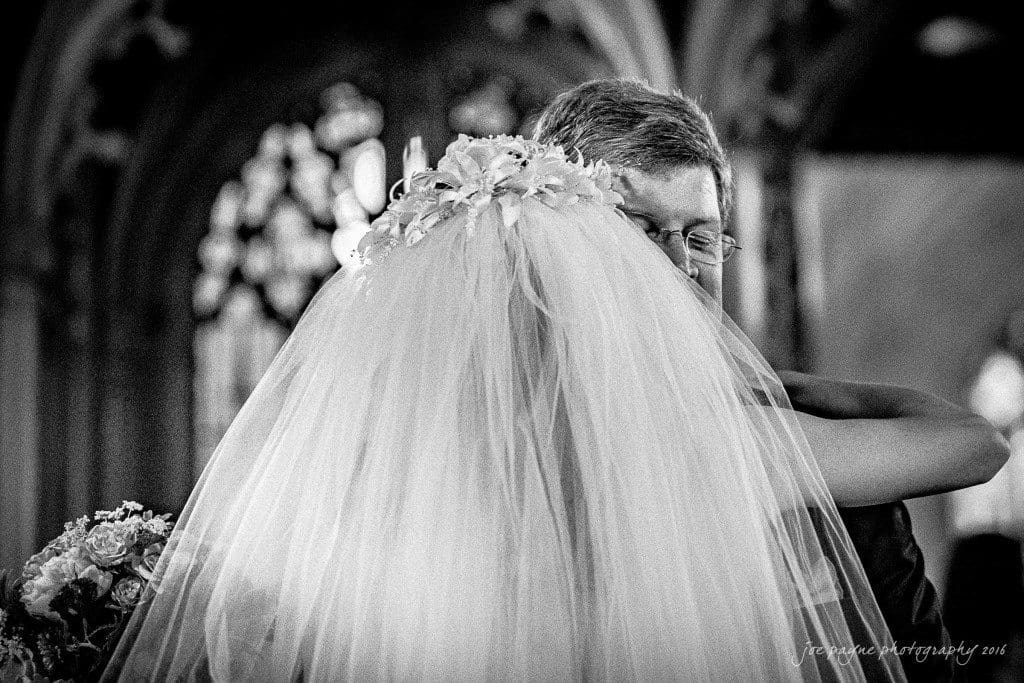 duke chapel & carolina inn wedding - sidney & andrew A groom embraces a bride whose back is facing the camera. Her veil and floral headpiece are prominent. They are standing in what appears to be a church, with arched windows creating a soft, blurred background. The scene is in black and white.
