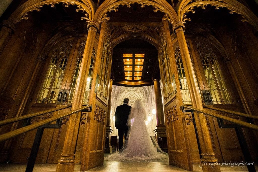 duke chapel & carolina inn wedding - sidney & andrew A bride and groom holding hands walk through ornate wooden doors into a grand cathedral, illuminated by soft, warm lighting. Intricate carvings and stained glass panels frame the entrance.