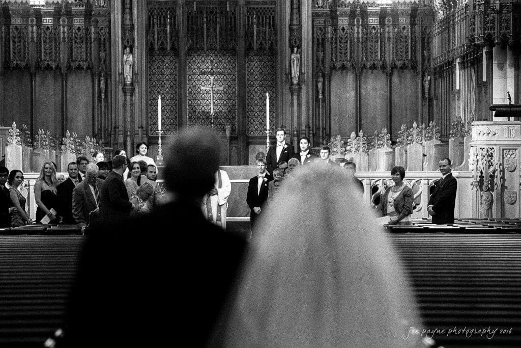 duke chapel & carolina inn wedding - sidney & andrew A black and white photo of a wedding ceremony in a grand church. The couple stands at the altar, surrounded by the wedding party and guests, with ornate architecture and intricate details in the background.