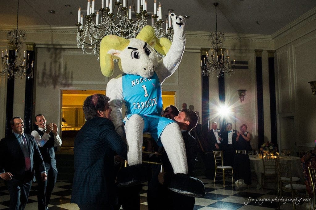 duke chapel & carolina inn wedding - sidney & andrew A group of people, dressed formally, are joyfully lifting a person in a ram mascot costume in a ballroom setting. The mascot is wearing a North Carolina jersey. Chandeliers and guests enjoying the event are visible in the background.