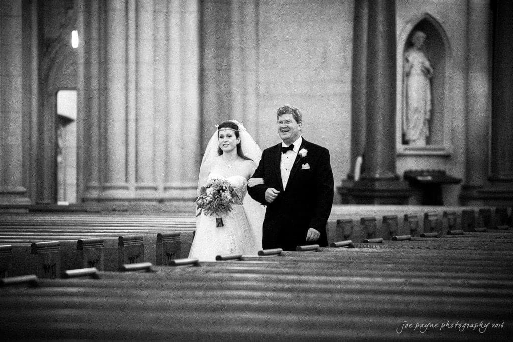 duke chapel & carolina inn wedding - sidney & andrew A bride in a white dress and veil walks arm in arm with a man in a tuxedo down the aisle of a cathedral, surrounded by empty pews. They both look forward, with statues and columns in the background.