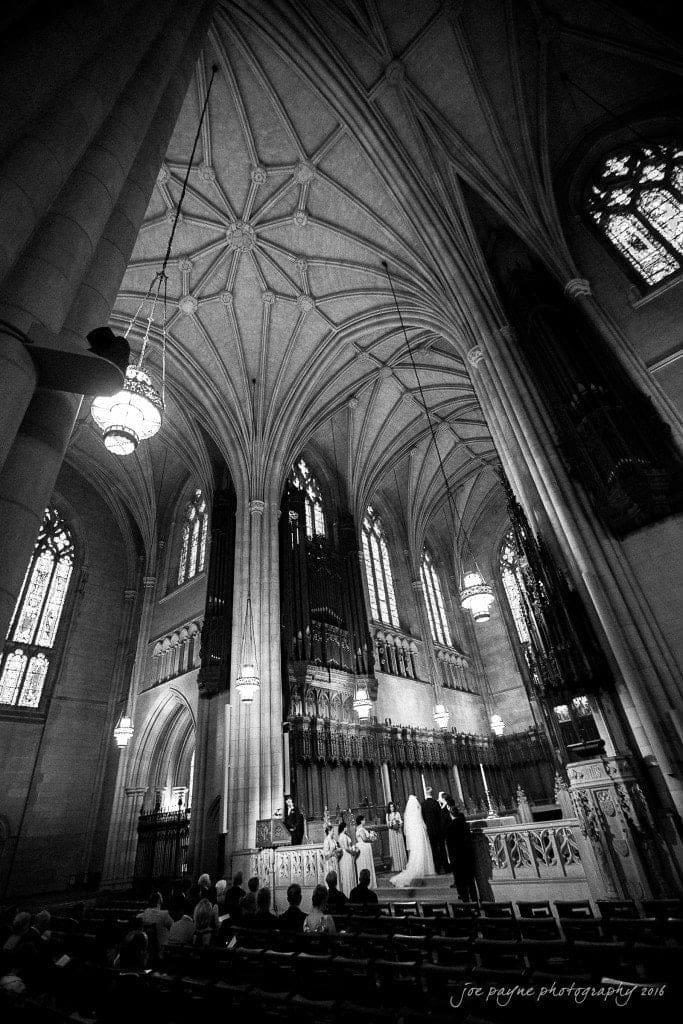 duke chapel & carolina inn wedding - sidney & andrew A black and white image of a grand church interior with high vaulted ceilings. A wedding ceremony is taking place at the front, with people seated in the pews. Tall arches and stained glass windows are visible in the background.