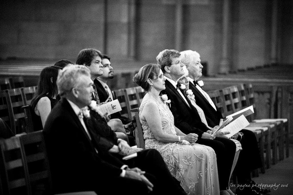 duke chapel & carolina inn wedding - sidney & andrew A group of people, dressed in formal attire, sit in wooden pews inside a church. Some are holding programs and wearing boutonnieres. The setting suggests they are attending a formal ceremony, such as a wedding or memorial.