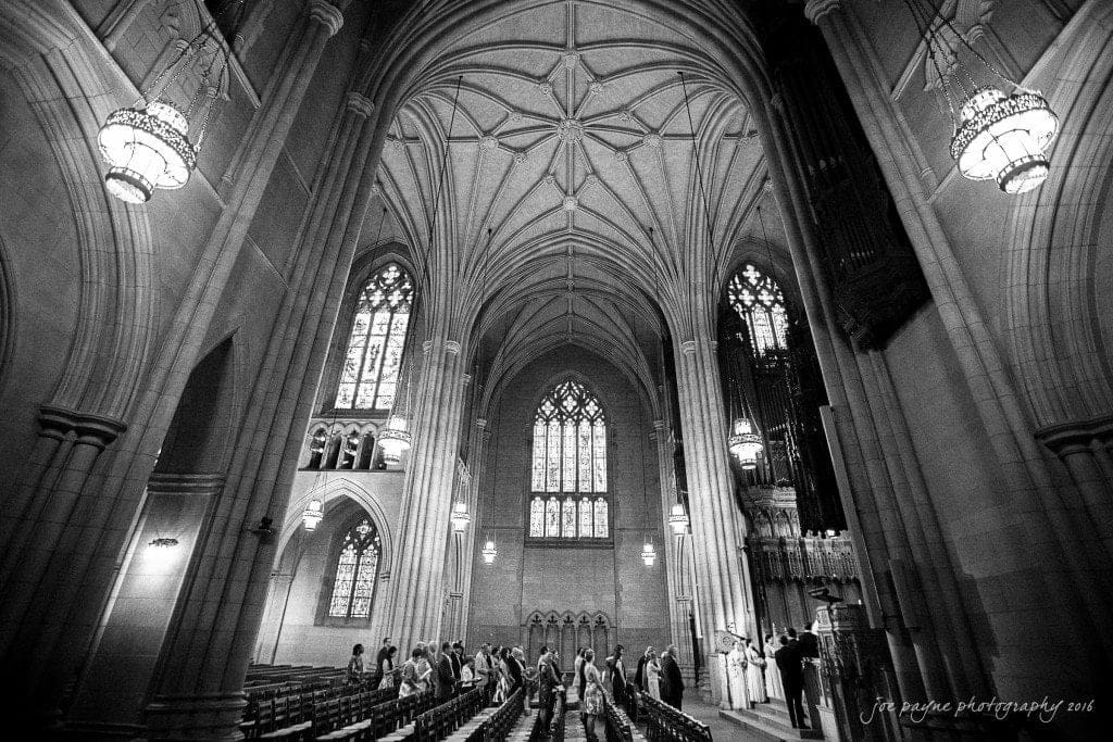 duke chapel & carolina inn wedding - sidney & andrew A black and white photo of a grand cathedral interior with high vaulted ceilings, stained glass windows, and chandeliers. A procession of people is walking down the aisle towards the altar.