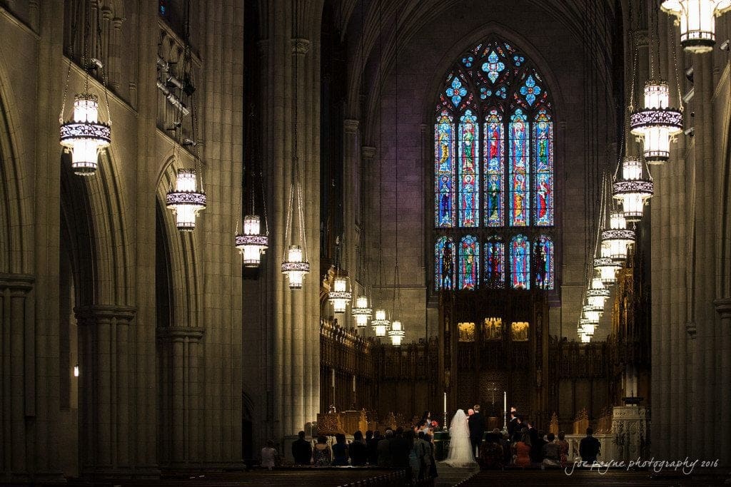 duke chapel & carolina inn wedding - sidney & andrew A wedding ceremony takes place inside a grand cathedral with high vaulted ceilings, ornate chandeliers, and colorful stained glass windows. The bride and groom stand at the altar, surrounded by guests seated in the pews.