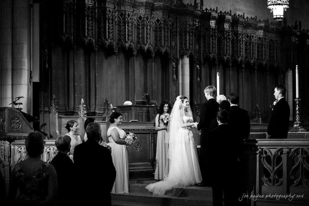 duke chapel & carolina inn wedding - sidney & andrew A bride and groom stand at the altar during a wedding ceremony in a church. Bridesmaids and groomsmen surround them, while guests look on. The setting features ornate wooden carvings and tall candles. The image is in black and white.
