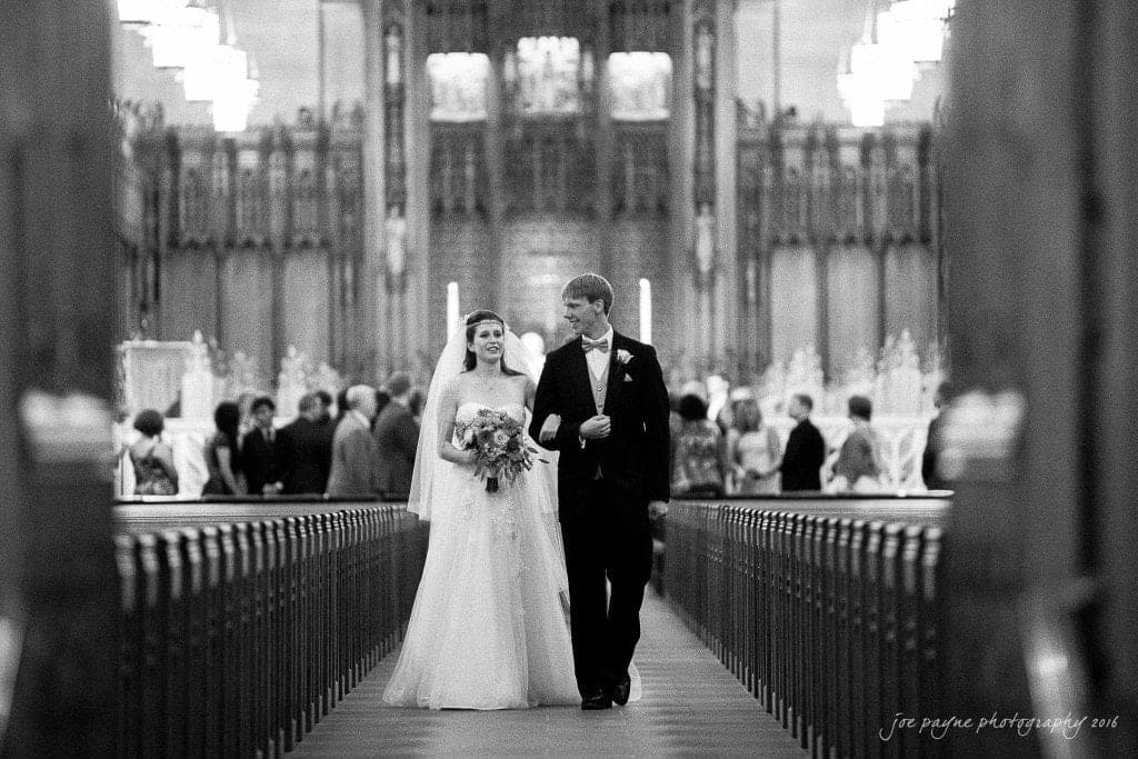 duke chapel & carolina inn wedding - sidney & andrew A bride and groom walk arm in arm down the aisle of a grand, ornate church. The scene is in black and white, with guests seated on either side. The churchs detailed architecture is visible in the background.