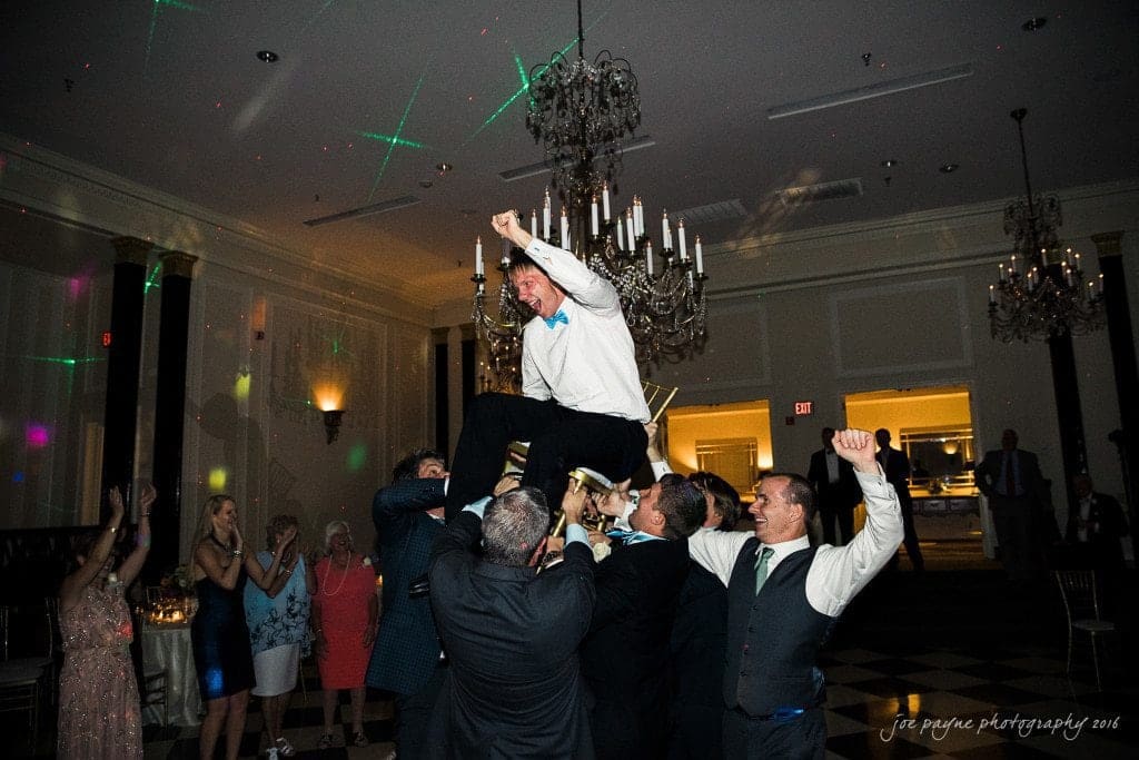 duke chapel & carolina inn wedding - sidney & andrew A joyful celebration at a wedding reception, with a man in a white shirt being lifted into the air by a group of people. Guests cheer and clap around the dance floor, while colorful lights shine across the room.