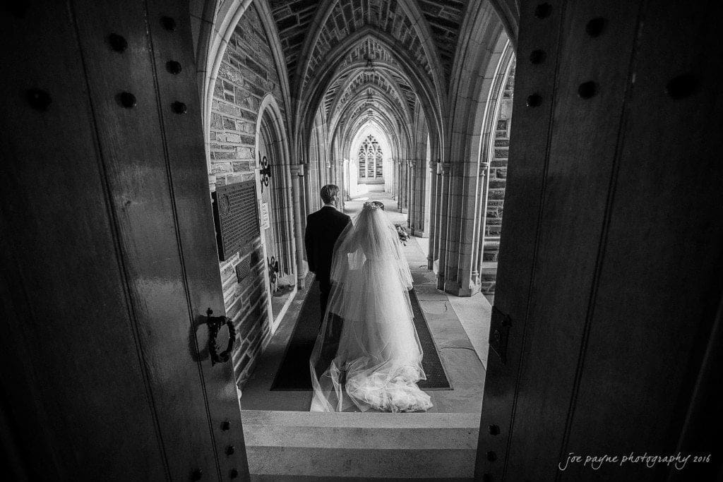 duke chapel & carolina inn wedding - sidney & andrew A bride and groom stand at the entrance of a historic stone church, framed by open wooden doors. The brides veil flows behind her, and the groom is beside her, both facing a beautifully ornate arched hallway. The scene is in black and white.