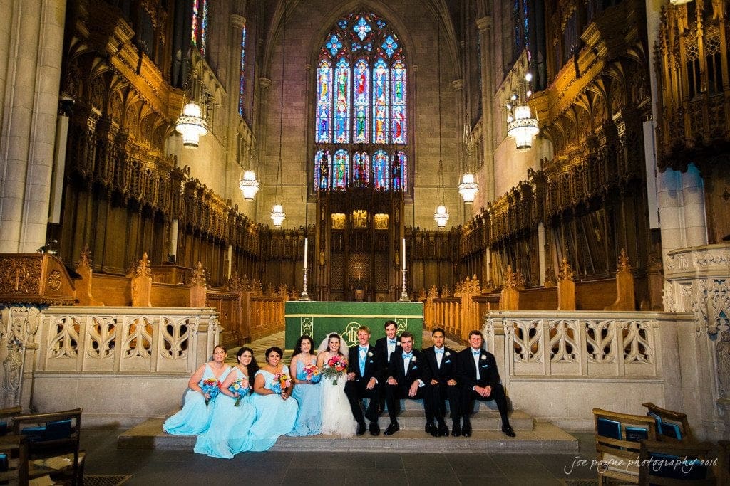 duke chapel & carolina inn wedding - sidney & andrew A bride and groom sit on a ledge inside a grand church with stained glass windows. They are surrounded by bridesmaids in light blue dresses and groomsmen in black suits. The space is ornate with high ceilings and decorative lights.