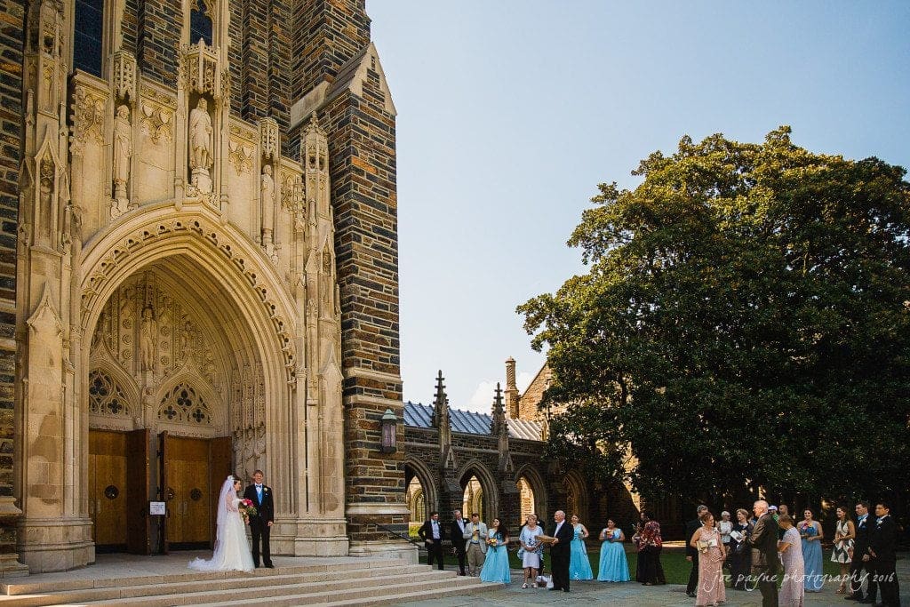duke chapel & carolina inn wedding - sidney & andrew A wedding ceremony outside a large, ornate stone church. The bride and groom stand by the entrance, while guests in formal attire gather around, watching and taking photos. A sunny day enhances the scene with a clear blue sky and lush greenery.