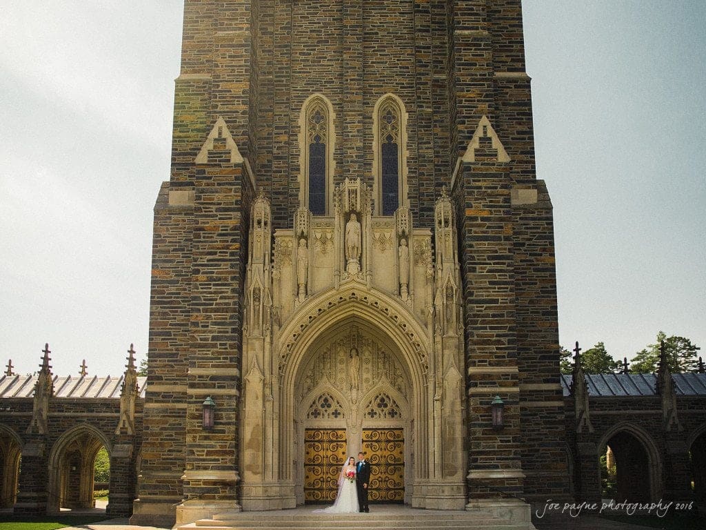 duke chapel & carolina inn wedding - sidney & andrew A couple stands in front of a large, ornate stone building with a tall archway and detailed carvings. The sky is clear, and the building has gothic architectural elements. The scene is bright and peaceful.