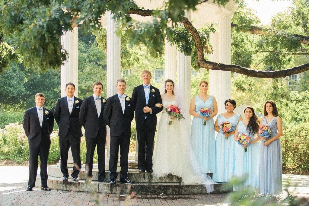 A bride and groom stand together with their wedding party under a white gazebo. The men wear black suits with bow ties, and the women wear light blue dresses while holding colorful bouquets. The setting is outdoors with lush greenery.
