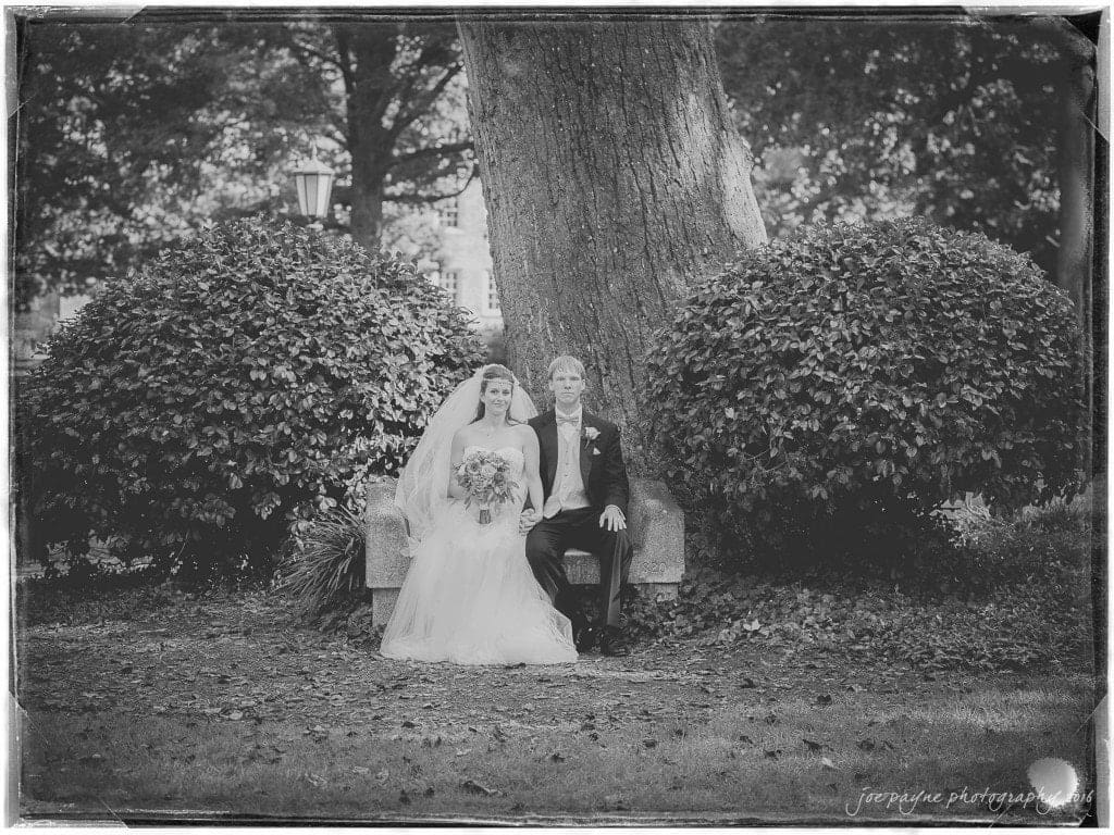 duke chapel & carolina inn wedding - sidney & andrew A bride and groom sit together on a stone bench under a large tree. The bride holds a bouquet and wears a veil, while the groom is in a suit. They are surrounded by lush greenery, creating a serene outdoor setting. The image is in black and white.